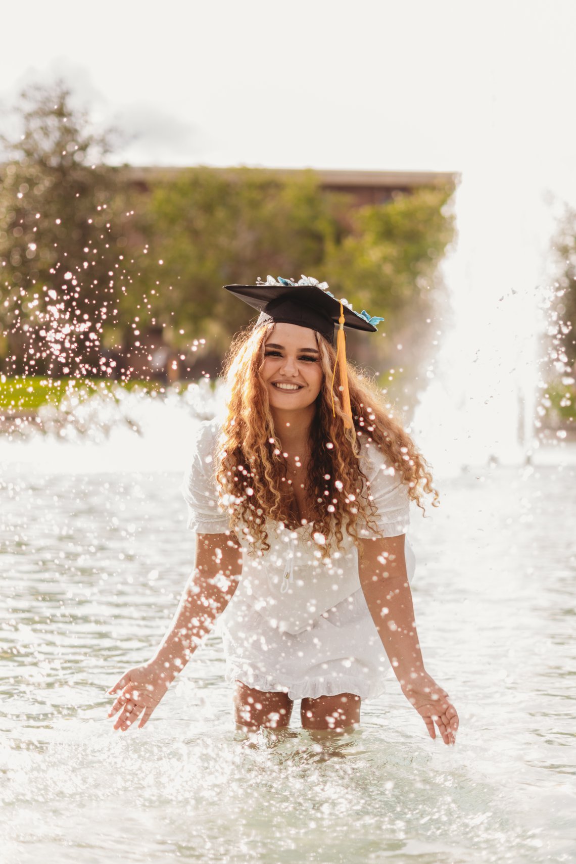 title A smiling young woman in a graduation cap and white dress stands in a fountain, with water splashing around her.